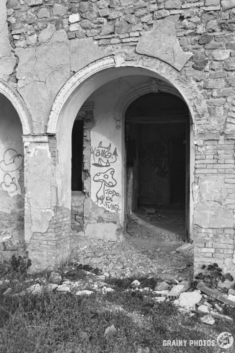 A monochrome image of an abandoned building with stone walls, featuring two arched doorways. One doorway reveals a wall with graffiti, depicting a whimsical character. The surroundings show rubble and overgrown vegetation.