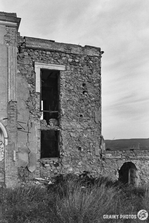 A partially collapsed stone wall of an old building, showing empty windows and overgrown grass in the foreground. The image captures a sense of decay and abandonment in a monochrome setting.