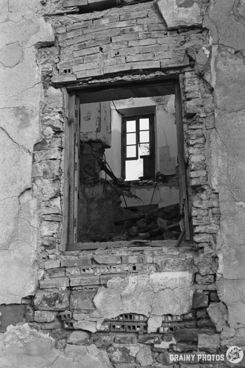 A monochrome image of a crumbling wall with a broken window, revealing the interior of a dilapidated room filled with debris and exposed architecture. The contrast highlights the textures of the brick and plaster.