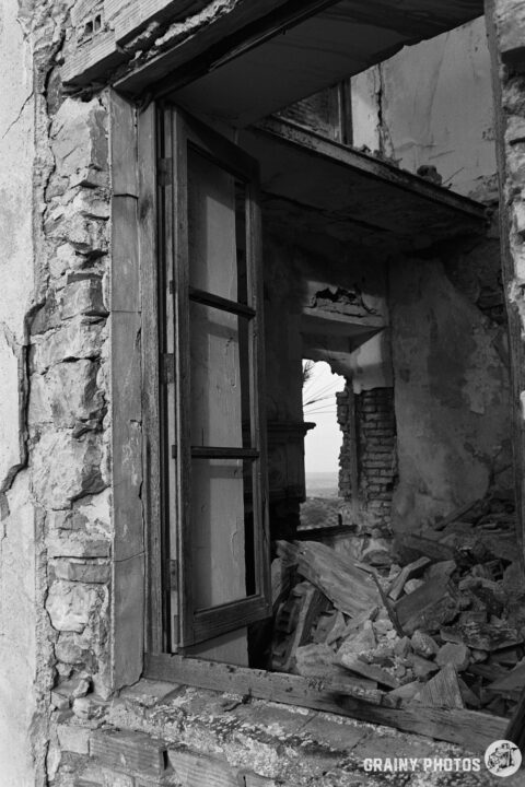 A partially collapsed building with exposed brick and debris visible through an open window. The scene captures a sense of decay and abandonment in a monochromatic tone.