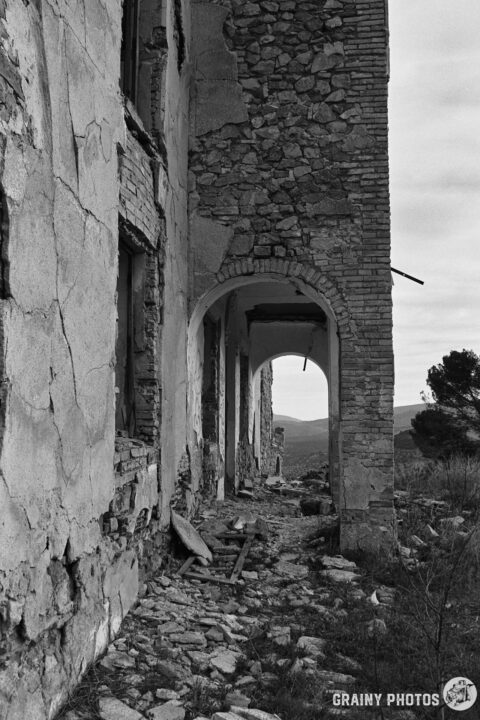 A weathered stone building with partially collapsed walls. An arched doorway leads into a derelict entrance porch, surrounded by rubble and vegetation, set against a cloudy sky and distant hills.
