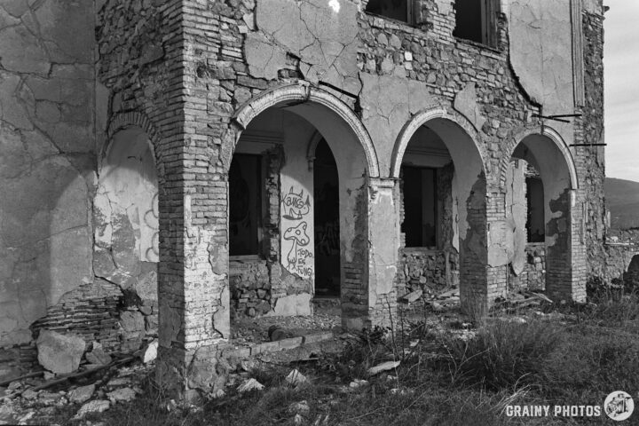 Black and white image of a dilapidated stone building with arches. The walls show peeling paint and graffiti, surrounded by overgrown grass and debris, reflecting a sense of abandonment and decay.