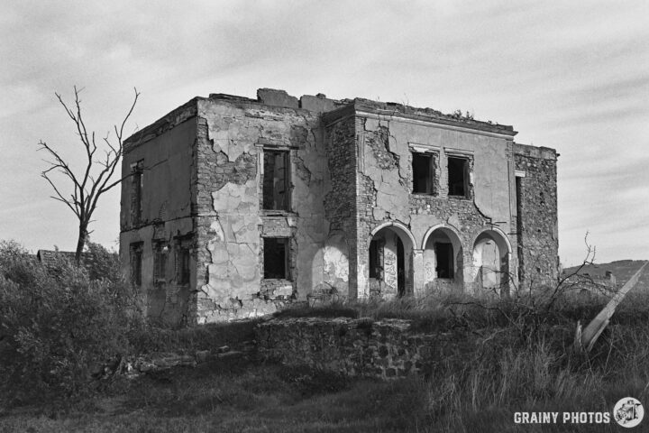 A dilapidated, two-story cortijo with crumbling walls and broken windows sits amid overgrown grass. A leafless tree stands nearby, contributing to the haunting atmosphere of abandonment in a black-and-white-style photograph.