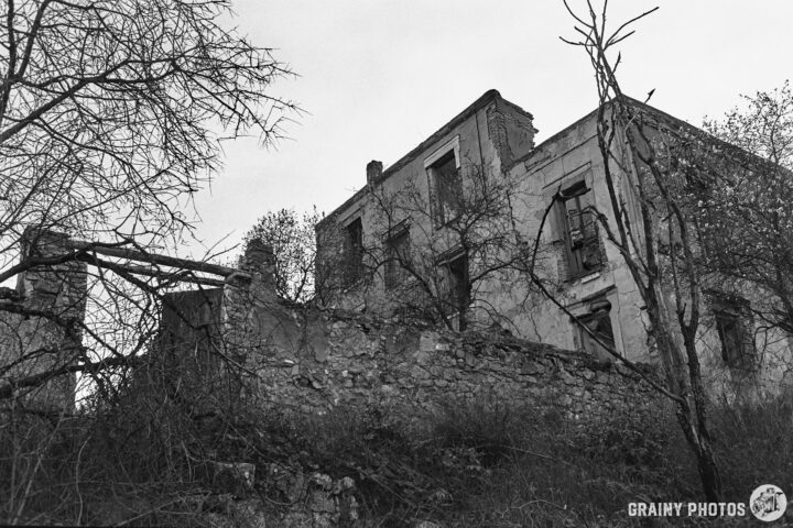 A dilapidated building with broken windows stands among overgrown trees, set against a cloudy sky. The structure's stone walls are partially crumbling, showcasing signs of decay in a forgotten landscape.