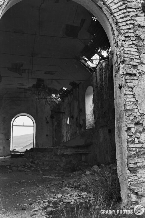 A desolate interior of an abandoned building, featuring crumbling brick walls, broken windows, and debris scattered on the floor, illuminated by natural light coming through a large arched window, creating a haunting yet captivating atmosphere.