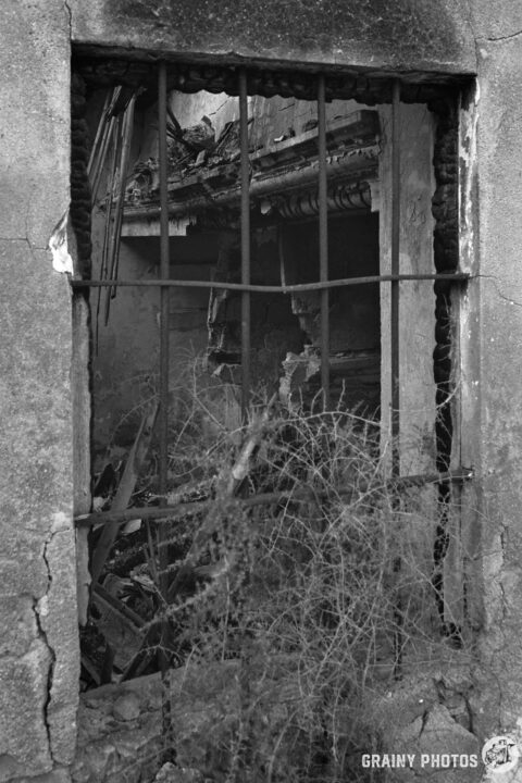 A dark, abandoned room is visible through a barred window, revealing collapsed structures and overgrown weeds, evoking a sense of decay and neglect in this black and white scene.