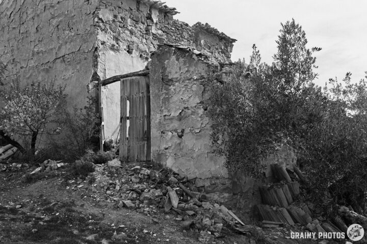 A weathered, partially collapsed building with a wooden door surrounded by overgrown vegetation and scattered stones, depicted in black and white.