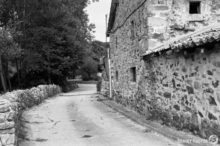 A quiet, winding rural road surrounded by stone walls and an aged stone building. Lush trees create a serene atmosphere in this black-and-white landscape, evoking a sense of tranquility and timelessness in nature.