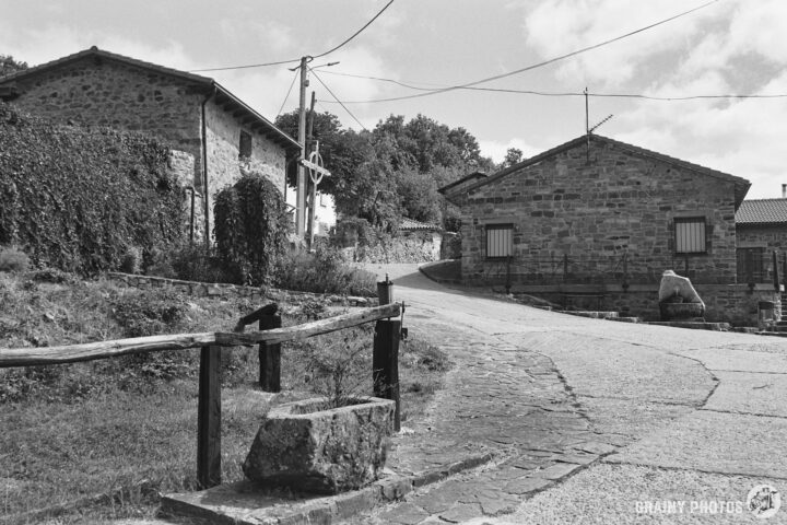 A picturesque village scene in Verdeña featuring rustic stone houses, a winding road, and greenery. The image is in black and white, capturing the tranquil charm of rural life under a part cloudy sky.