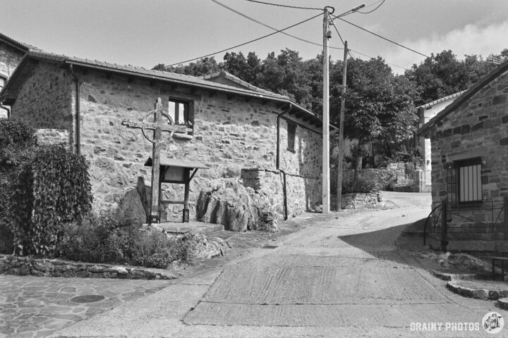 A rustic stone house stands at the edge of a winding road in Verdeña. A wooden cross is nearby, surrounded by greenery and rocks, with trees in the background. The scene captures a serene, historical atmosphere.