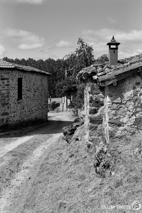 A winding dirt road leads through Verdeña, flanked by rustic stone houses with tiled roofs. A lamp post adds charm to the picturesque scene under a partly cloudy sky.