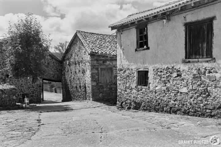 Black and white photograph of a rustic village scene with stone houses, a narrow path, and a cloudy sky. The buildings feature weathered facades and wooden windows, evoking a sense of history and charm in a tranquil rural setting.