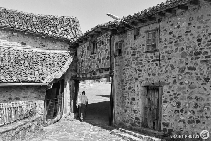 A person walking under a tinao between two rustic stone buildings with tiled roofs, set in a quaint village scene. The image is in black and white, emphasizing the textures and historical architecture of the surroundings.
