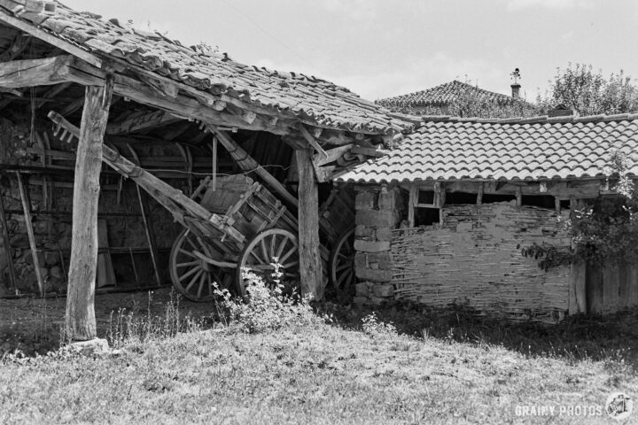 A rustic scene featuring an old wooden barn with a traditional cart beside it. The structure has a tiled roof, and the surrounding area is overgrown with grass, capturing the charm of rural architecture in black and white.