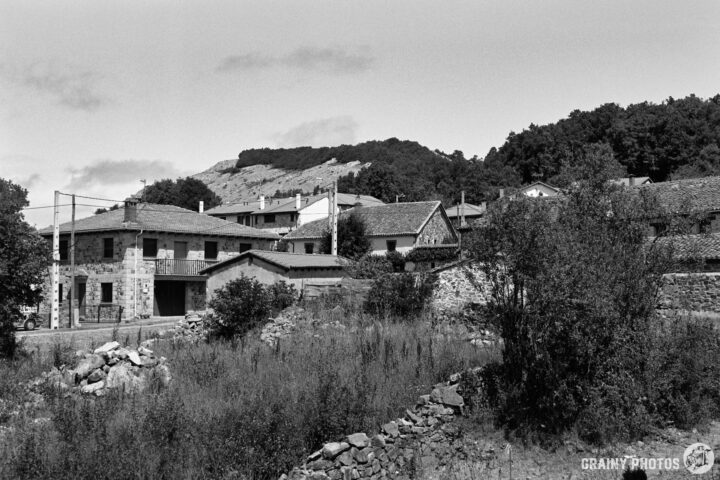 A serene black-and-white landscape featuring quaint stone houses nestled among greenery, with a backdrop of rolling hills and trees under a cloudy sky. The scene evokes a sense of tranquility and rural charm.