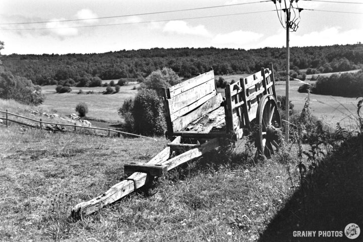 A rustic wooden cart rests in a field, surrounded by trees and rolling hills. The scene reflects a peaceful rural landscape under a bright sky with scattered clouds.