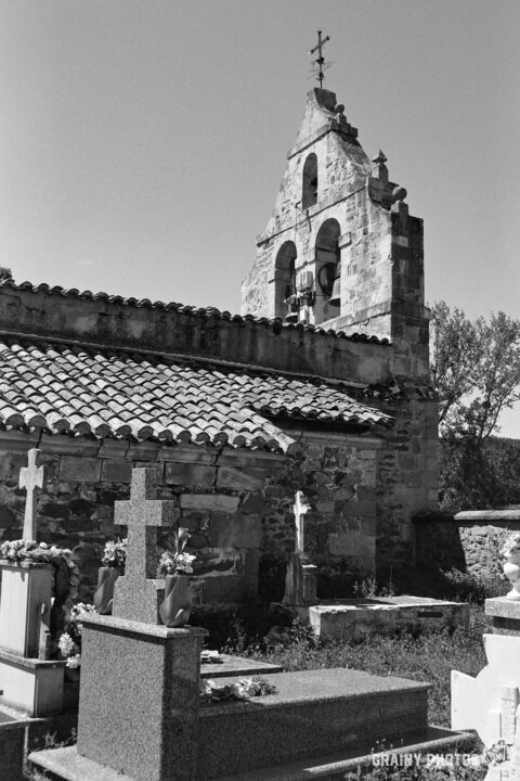 A black-and-white image of San Miguel Church with a bell tower in Verdeña, surrounded by graves marked by simple crosses. Lush greenery partially frames the scene under a clear sky, creating a serene and reflective atmosphere.