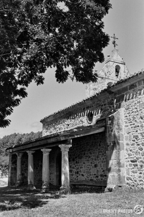 Black and white photograph of San Miguel Church in Verdeña featuring a bell tower. The structure has a covered porch supported by columns, surrounded by lush green trees, creating a serene rural atmosphere. The scene captures historical architecture and nature harmoniously.