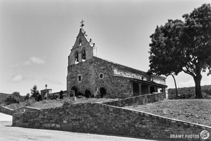 San Miguel Church in Verdeña with a bell tower, set against a clear sky. The building features decorative elements and is surrounded by trees and greenery, showcasing a serene landscape. The scene captures a timeless architectural beauty.