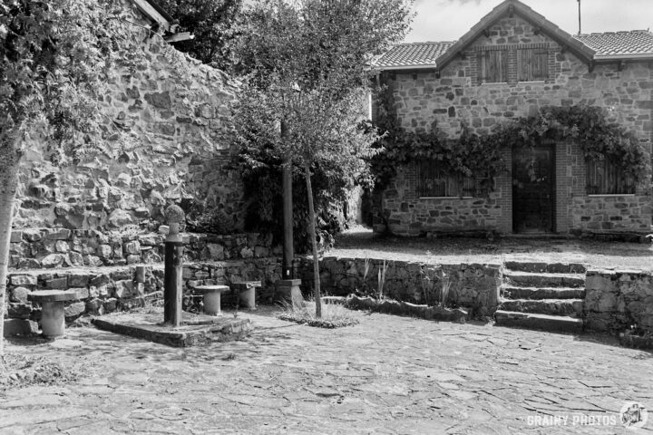 A rustic stone courtyard features an old water pump, simple stone steps, and a charming stone building with a sloped roof, surrounded by greenery. The scene is captured in black and white, adding a vintage atmosphere.