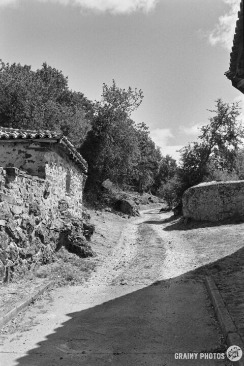A winding dirt path leads through a rural landscape, bordered by stone walls and trees. The scene captures a tranquil atmosphere of nature and rustic architecture under a partly cloudy sky.