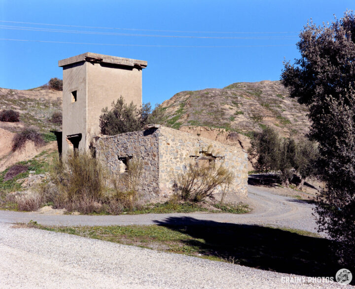 A weathered stone building and an adjacent tower stand on a gravel path surrounded by hills and brush, under a clear blue sky. The landscape features arid terrain with sparse vegetation.