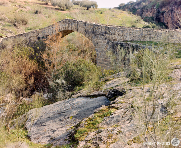 A historic stone Roman Bridge arches over a stream, surrounded by lush greenery and rocky terrain, showcasing natural beauty and architectural craftsmanship in a serene outdoor setting.