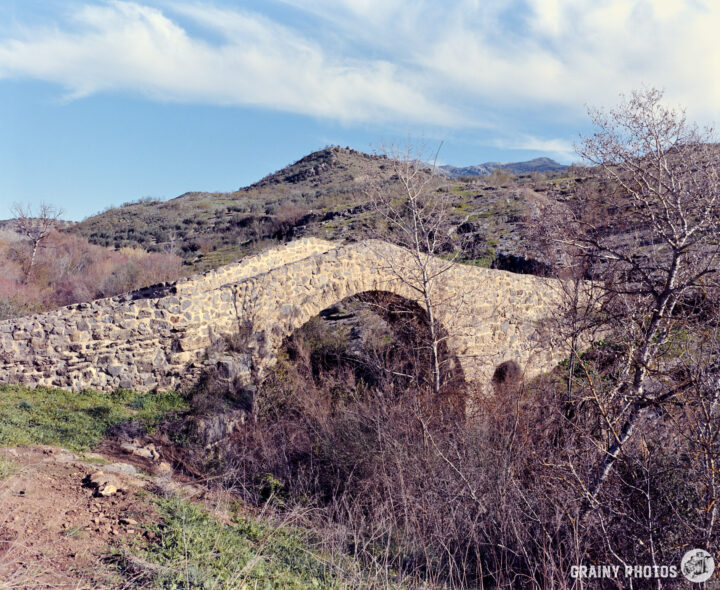 A historic Roman bridge arches over a lush landscape adorned with sparse trees and rolling hills under a blue sky with wispy clouds. The scene captures the beauty of nature and architectural heritage.
