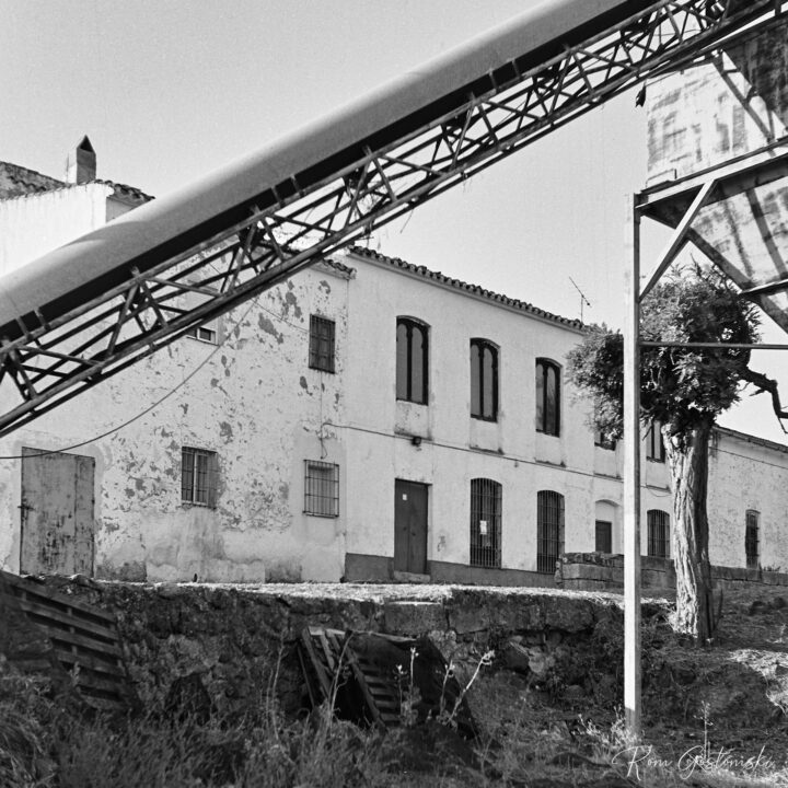 A black and white photograph of a rustic, weathered cortijo with barred windows, surrounded by overgrown vegetation. A large industrial structure with a conveyor belt and hopper is visible in the foreground.