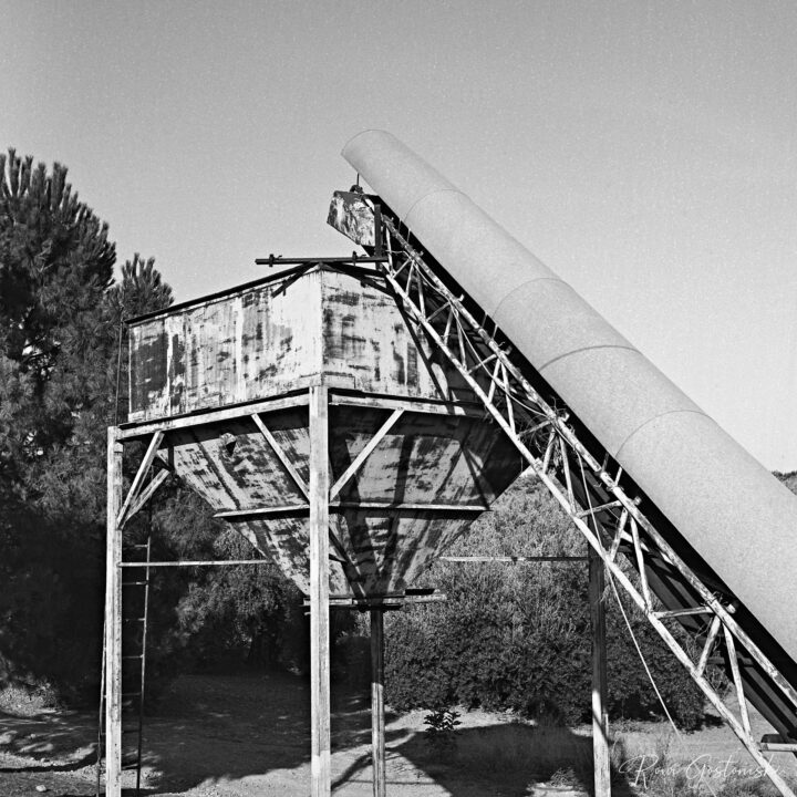 A weathered industrial silo and hopper with a large conveyor with cylindrical cover, surrounded by trees under a clear sky, captured in black and white.