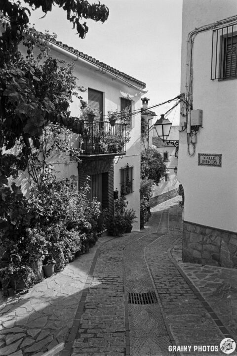 A narrow, winding cobblestone street lined with white buildings adorned with balconies and greenery. A vintage street lamp hangs on a wall, while a sign reads "Calle Estación.” The scene captures a quaint, historical charm in black and white.
