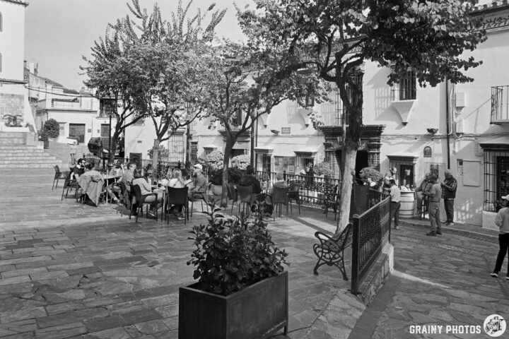 A bustling outdoor plaza surrounded by trees, featuring tables filled with diners enjoying their drinks and meals. In the background, charming white buildings and a few pedestrians create a lively atmosphere in this picturesque setting.