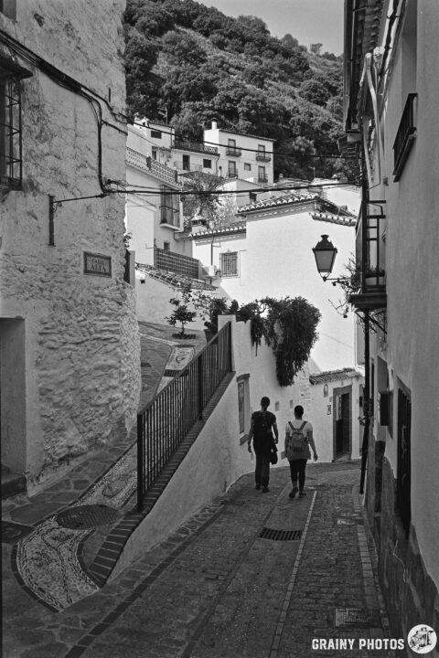 A narrow, winding street in Benelauría, flanked by whitewashed buildings with traditional architecture. Two people stroll down the cobblestone path, surrounded by greenery on the hillside above. A vintage street lamp adds charm to the scene.