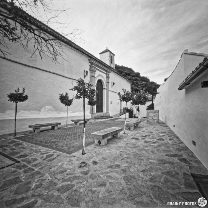 A black and white photo of a quiet, narrow plaza lined with small trees and stone benches. On the left is the Santo Domingo de Guzmán church with a bell tower. Soft, overcast sky adds to the serene atmosphere.