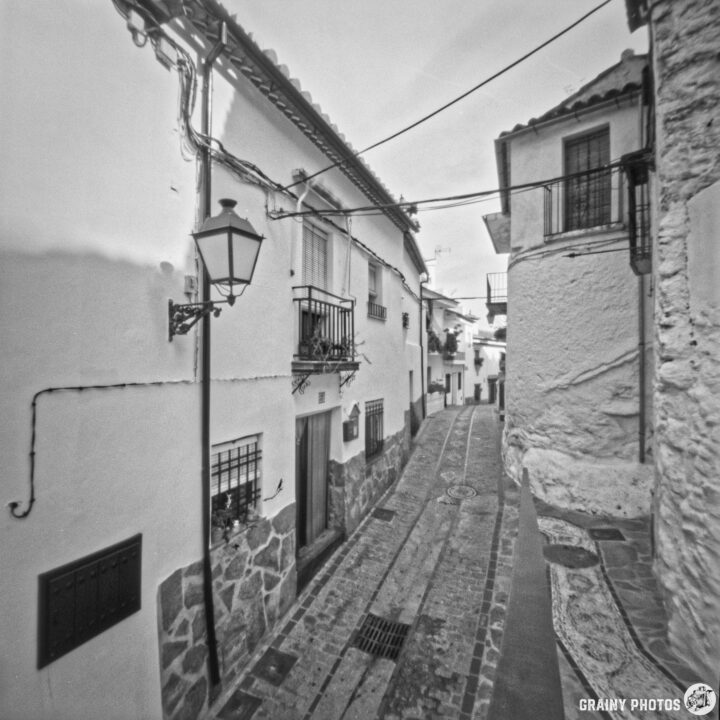 A narrow cobblestone street winds between traditional whitewashed buildings with balconies and vintage lanterns, creating a charming atmosphere in Benelauría. The scene captures a mix of urban and rustic elements in black and white.