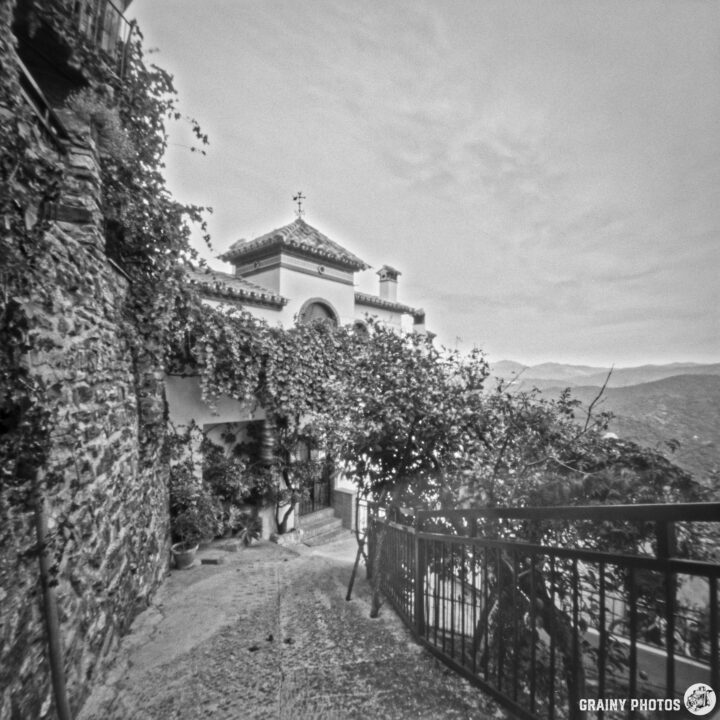 A narrow pathway leads to a charming old building covered in vines, surrounded by scenic hills in black and white. The pathway features stone walls and railings, highlighting the tranquil and rustic atmosphere of the area.
