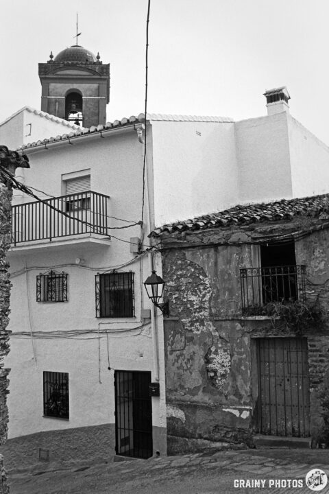 A black-and-white image of a quaint street scene featuring historic buildings. The architecture includes a mix of whitewashed exteriors and textured stone walls, with a prominent bell tower in the background and decorative window grilles.
