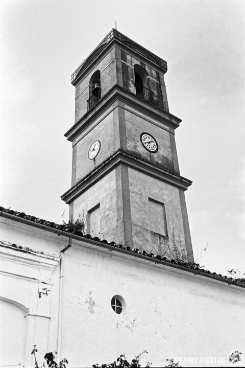 A black and white photograph of a historic church tower with clock and a bell, featuring a weathered exterior, situated against a clear sky above a white church wall.
