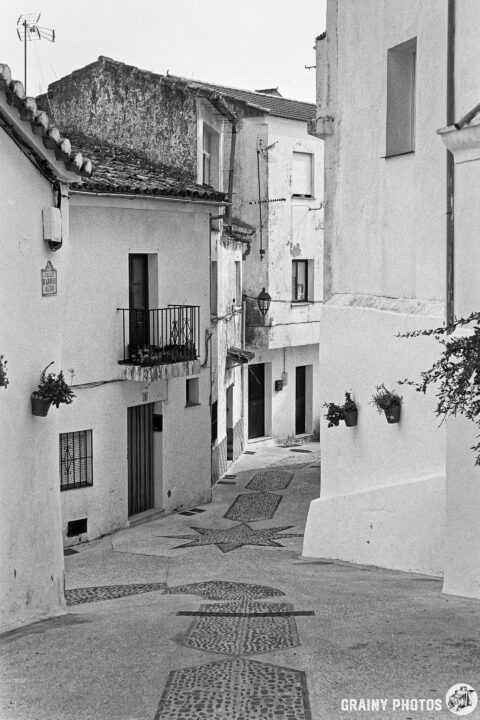 A narrow, cobblestone street meanders through Algatocín, lined with houses featuring balconies and flower pots. The scene captures the quiet atmosphere of a historic area, evoking a sense of nostalgia and tranquility.