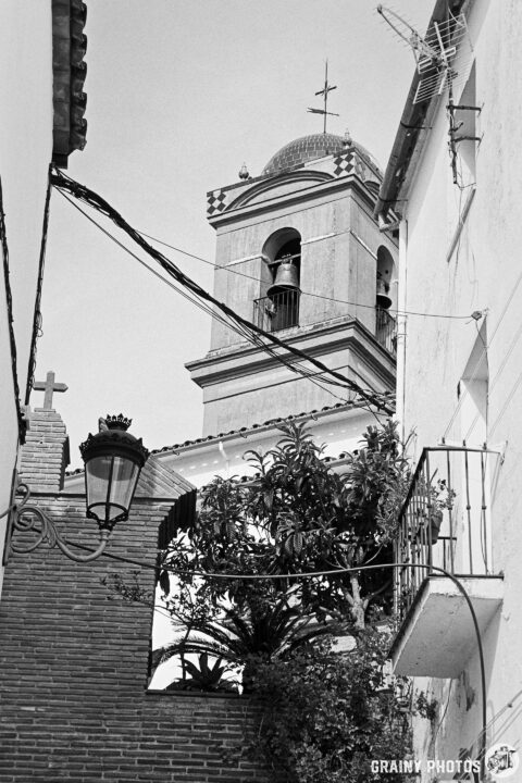A charming black and white view of a narrow street featuring a bell tower, adorned with wires and a wall lantern, framed by lush greenery and architectural details of nearby buildings.