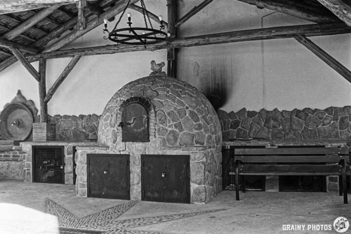 An indoor space featuring a comunal stone bread oven with a domed top, set against a rustic wooden structure. The walls are adorned with stone, and there’s a bench for seating. A chandelier hangs from the ceiling, adding to the charm of the setting.