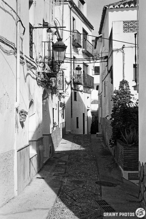 A narrow, cobblestone alleyway lined with whitewashed buildings, featuring balconies and street lamps. Sunlight casts shadows, creating a tranquil atmosphere in this charming urban setting.