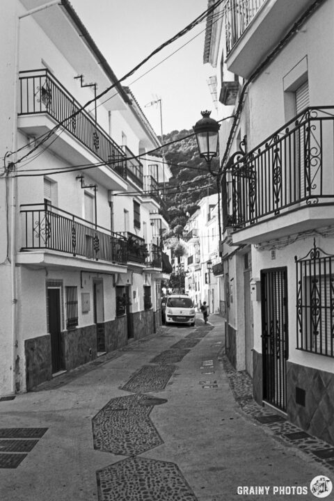 A narrow, cobblestone street in Algatocín features white buildings with balconies and ornate railings. A van is parked nearby, and steep hills are visible in the background, suggesting a picturesque and peaceful atmosphere.