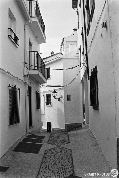 A narrow, winding alleyway lined with white buildings, featuring balconies and a street lamp, creating a charming and serene atmosphere. The cobblestone path adds texture to the scene, typical of quaint Spanish village architecture.