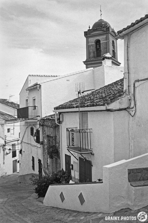 A narrow, winding street in Algatocín framed by whitewashed houses with balconies, leading up to a bell tower. The scene is captured in black and white, highlighting the charming architecture and cobblestone details.