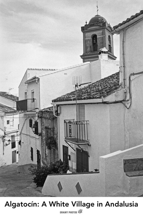 Analog travel photo of a narrow, winding street in Algatocín framed by whitewashed houses with balconies, with a bell tower in the distance. The scene is captured in black and white, highlighting the charming architecture and cobblestone details.