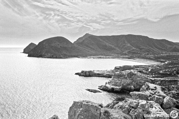 A black and white photograph depicting a coastal landscape with rugged cliffs and mountains in the background, overlooking a calm sea. The sky features soft clouds, creating a serene and dramatic atmosphere.