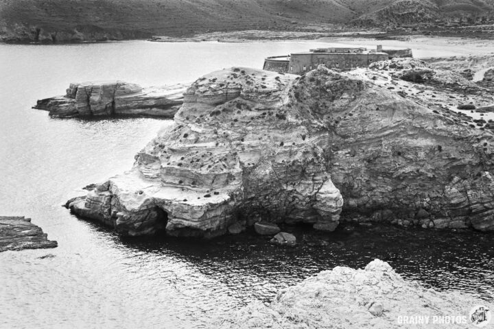 A black and white image of rocky cliffs by a calm sea, featuring a small fort at the top of the cliff, surrounded by rugged terrain and water reflecting the landscape.
