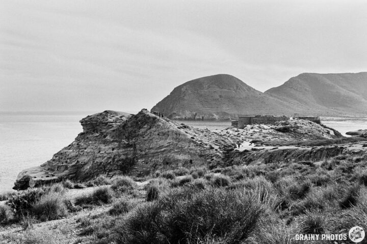 A rugged coastline featuring prominent rock formations, Castillo de San Ramón and rolling hills, with calm waters in the background under a cloudy sky. The scene reflects a serene, natural landscape in black and white.