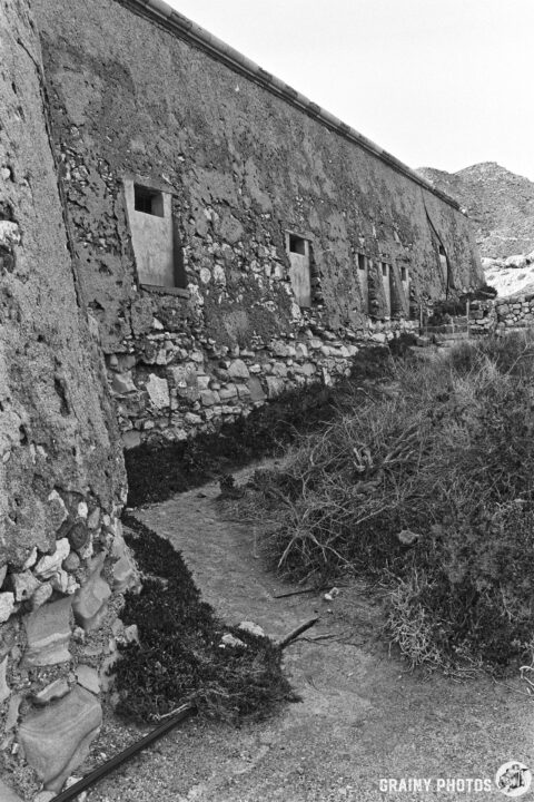 The rear of Castillo de San Ramón with partially closed windows, set against a backdrop of rocky terrain and sparse vegetation, captured in black and white. A narrow pathway leads along the structure, suggesting an abandoned or historical site.